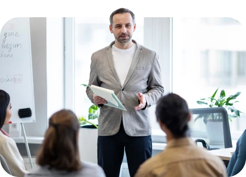 Homem de terno cinza e camisa branca fazendo palestra para um grupo de pessoas em ambiente de escritório moderno e bem iluminado.